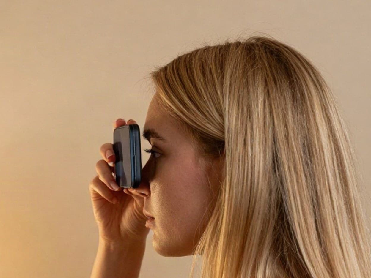 Side close-up at the mirror: the back of the phone resting against her brow, the lens over her eye